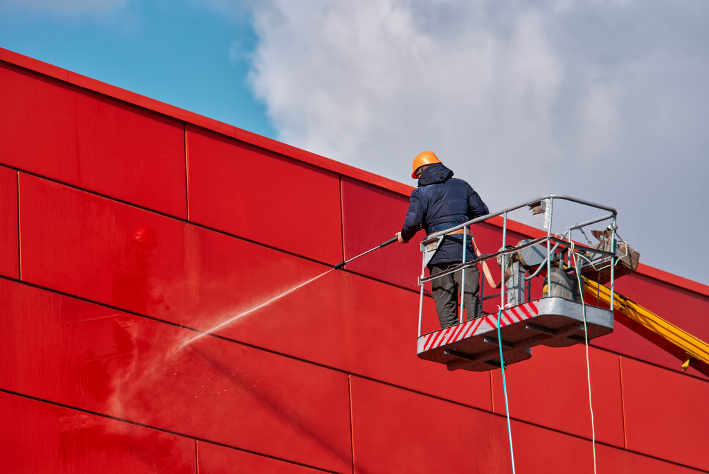 A person on a crane pressure washing a red wall of a modern commercial building
